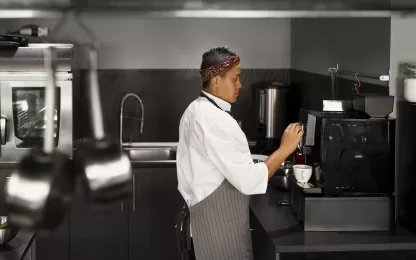 Chef operando máquina de café em cozinha profissional enquanto prepara xícaras, próximo a área equipada para lavagem de copos e xícaras.