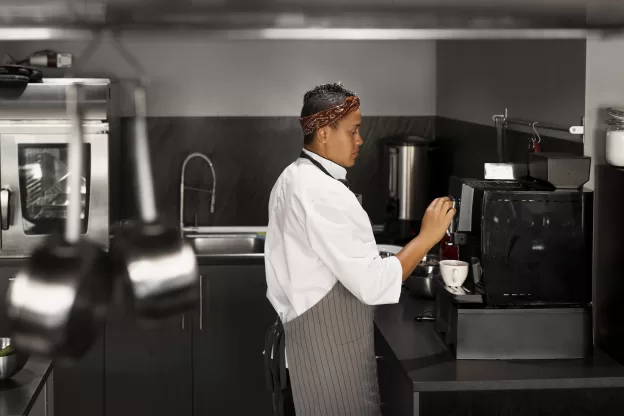Chef operando máquina de café em cozinha profissional enquanto prepara xícaras, próximo a área equipada para lavagem de copos e xícaras.