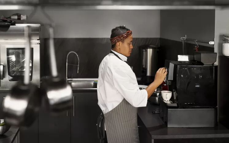 Chef operando máquina de café em cozinha profissional enquanto prepara xícaras, próximo a área equipada para lavagem de copos e xícaras.
